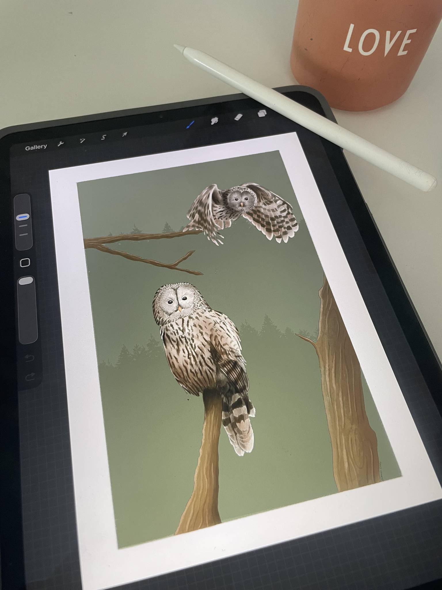 A tablet showing a digital illustration of two owls perched on branches, with a white stylus resting beside it and part of a terracotta candle in the top-left corner.